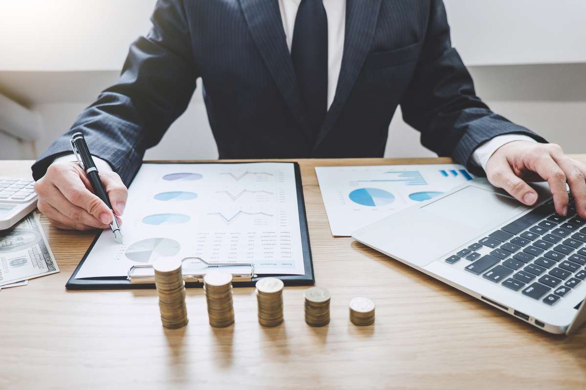 Businessman writing making report with stacked coins arranged at office desk and many document data graph, business concept (R) (S)