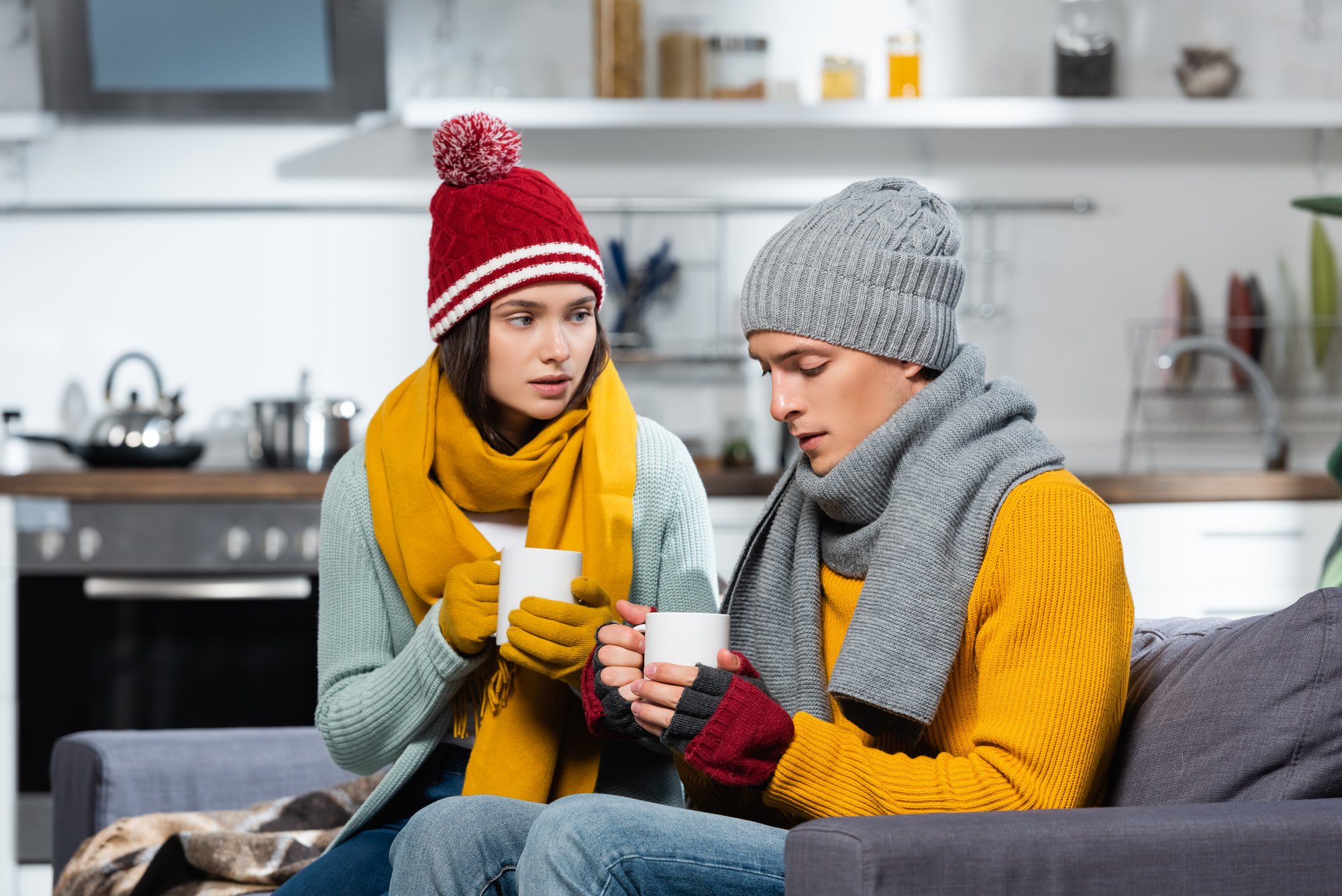 Freezing couple in knitted hats, scarfs and gloves holding cups of warming beverage in cold kitchen