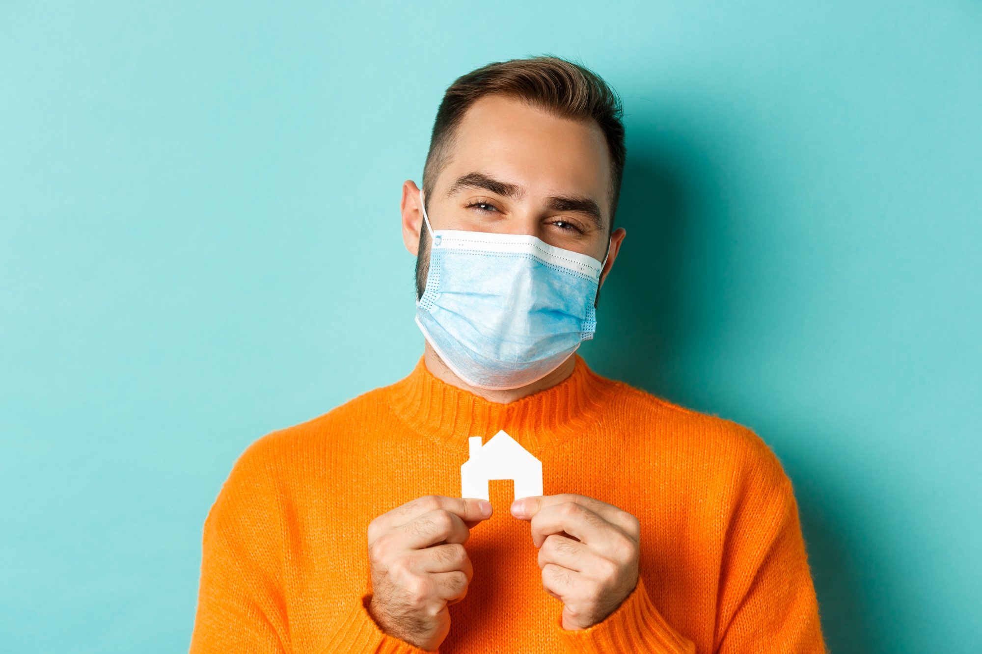 Real estate and coronavirus pandemic. Close-up of adult man in medical mask holding small paper house maket and smiling, searching for apartment