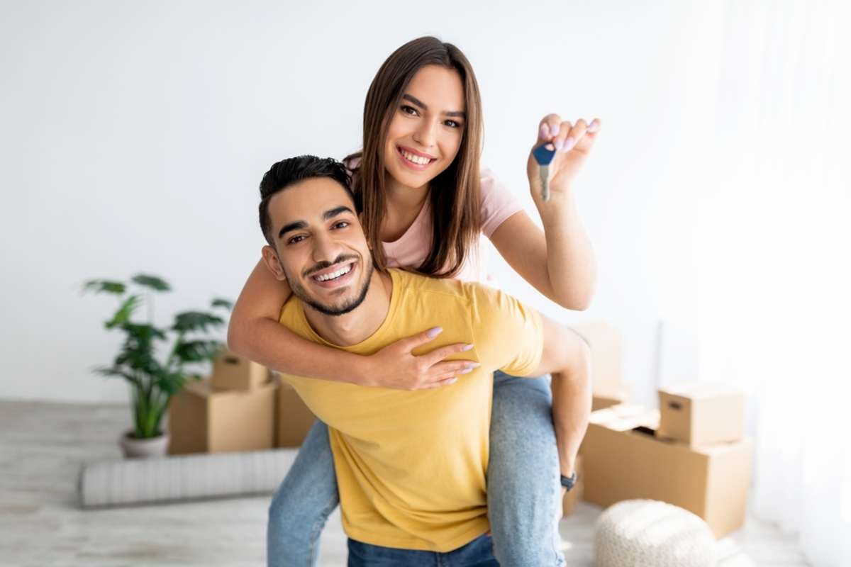 Cheerful Caucasian lady getting piggyback ride from her Arab boyfriend, showing house key indoors