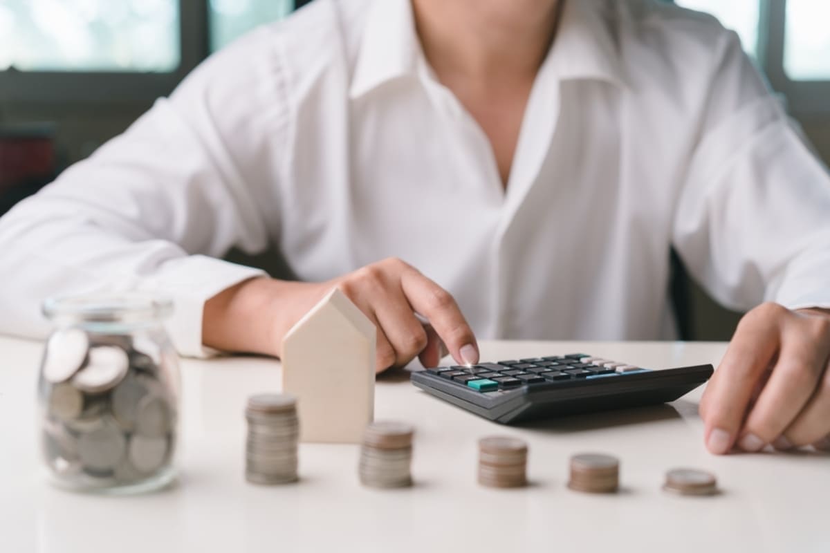 Person using a calculator next to stacks of coins and a model house, average property management fees concept