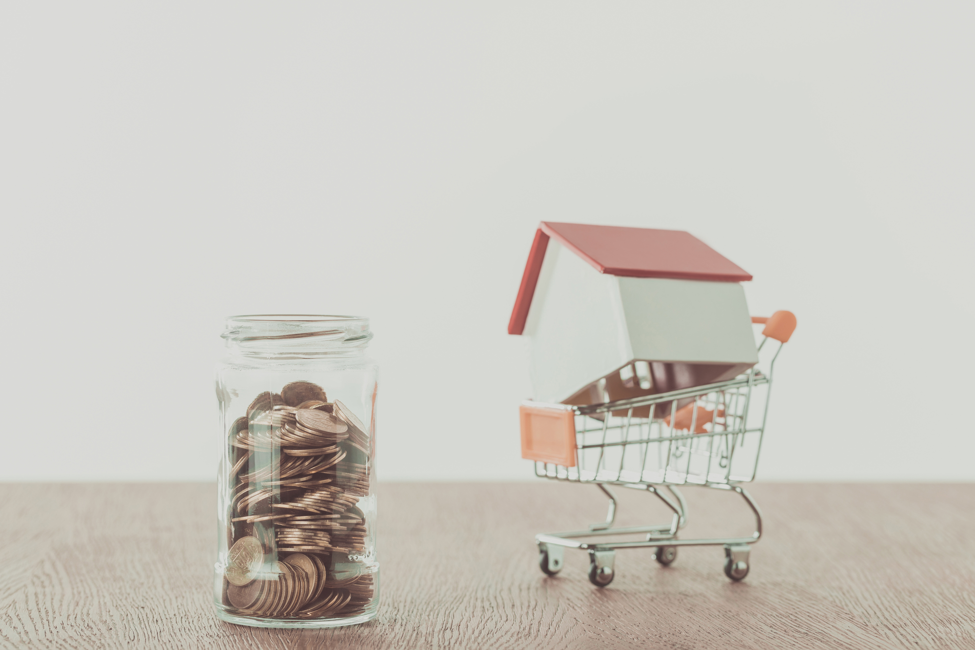 Small wooden house on supermarket cart, jar of coins on wooden table, saving concept